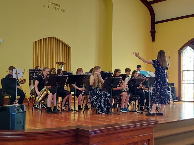 Group of youth in concert band playing their instruments on stage for a public performance.