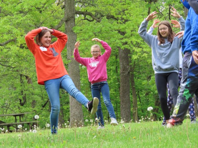 Group of youth playing a drama game standing on one foot with their hands on their heads.